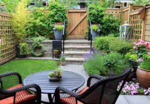 Enclosed backyard patio space with lots of green bushes and flowers and black wicker table and chair set